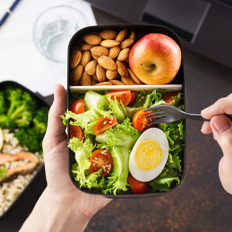 Photo of a man's hands holding a fork to eat from a bento box filled with salad, nuts, and an apple.