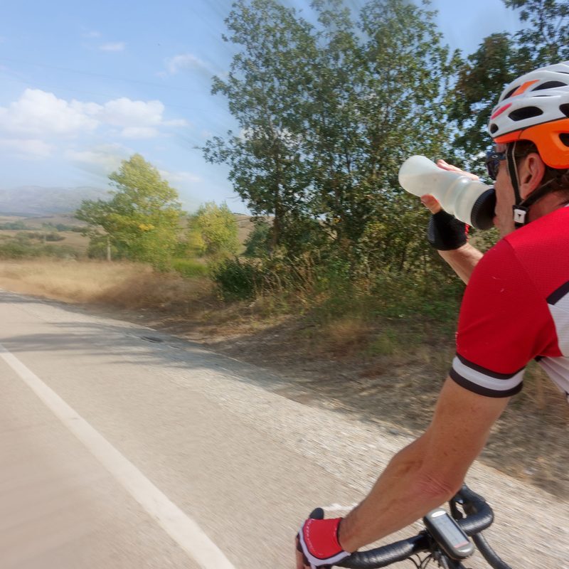 A biker drinking an electrolyte beverage from his water bottle while cycling on the street.