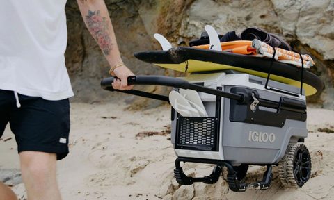 Man pulling a wheeled Igloo cooler over sand at the beach.