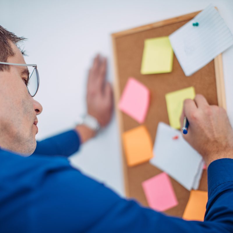 A man with glasses on writing reminders on a sticky note placed on a corkboard.