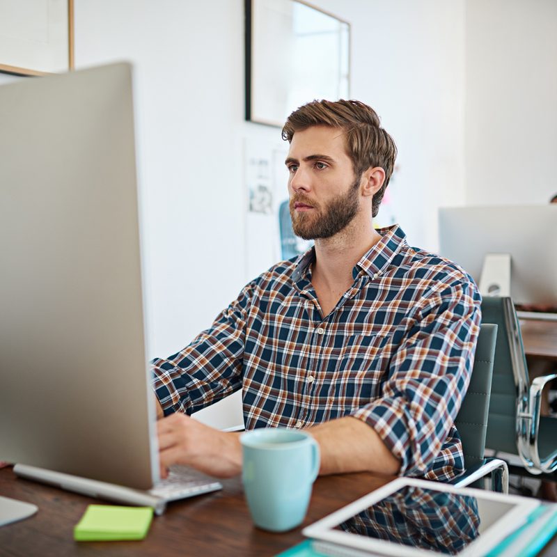 A young and bearded man wearing a plaid shirt at the office while typing on his Mac.