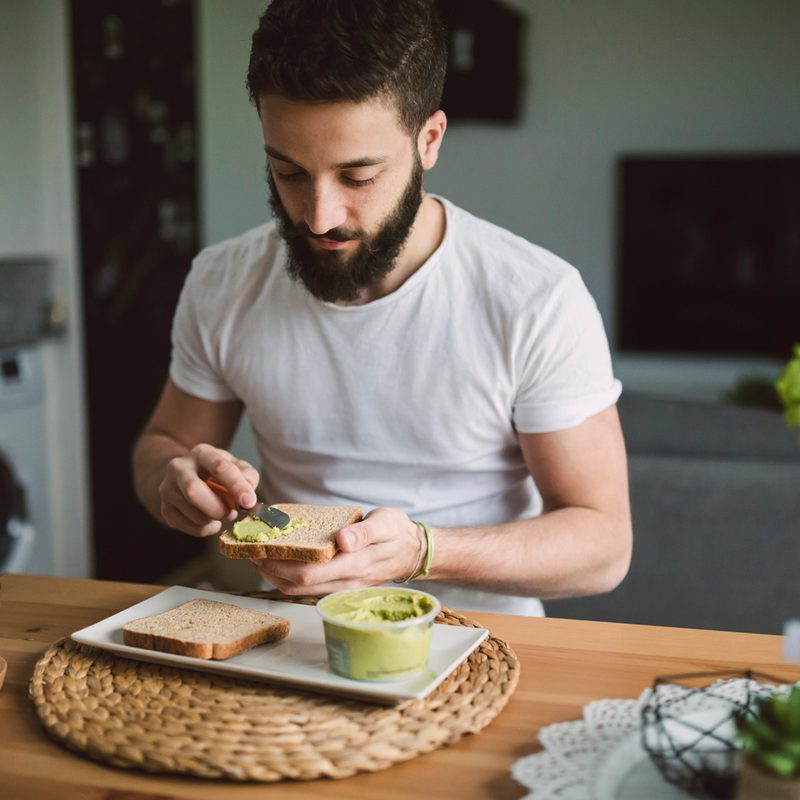 A bearded man preparing guacamole and toast on his wooden kitchen table.