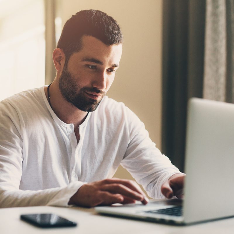 A man with a buzz-cut and beard using his laptop with his phone on the side.