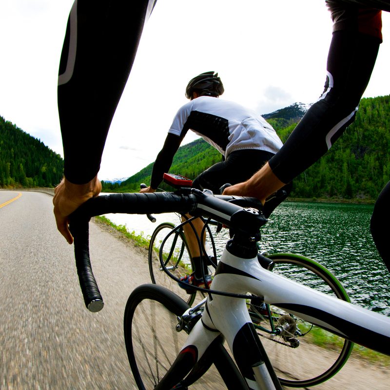 A man's hands holding the pedal of his bicycle as another male cyclist overtakes him on a road by a body of water.