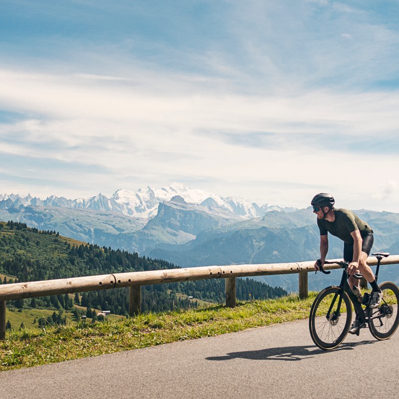 A man in helmet riding a bike on a road with railings with a view of a mountain and forest in the background.