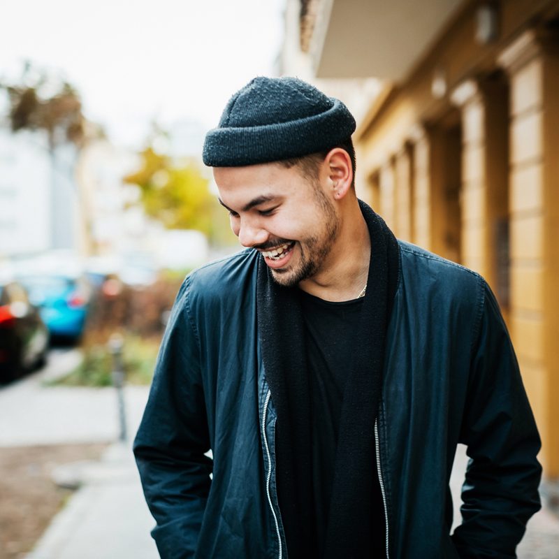 A smiling man wearing a beanie and jacket as he takes a stroll around the street.