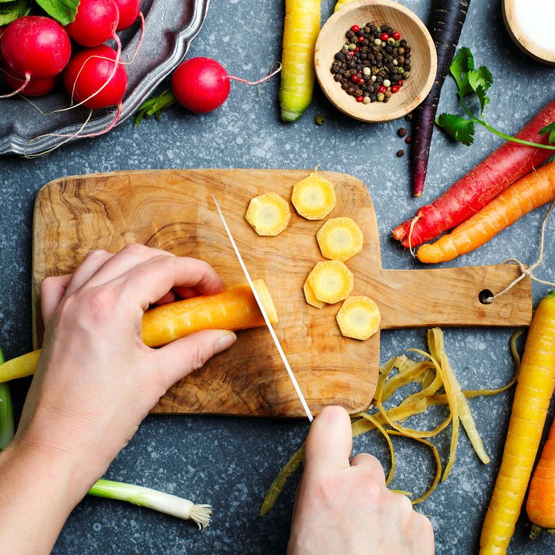 Cutting carrots on wooden board and fresh spring vegetables for vegetarian cooking on the table, top view.