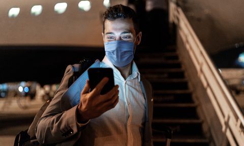 what the covid vaccine means for travel businessman using mobile phone at airport protective mask