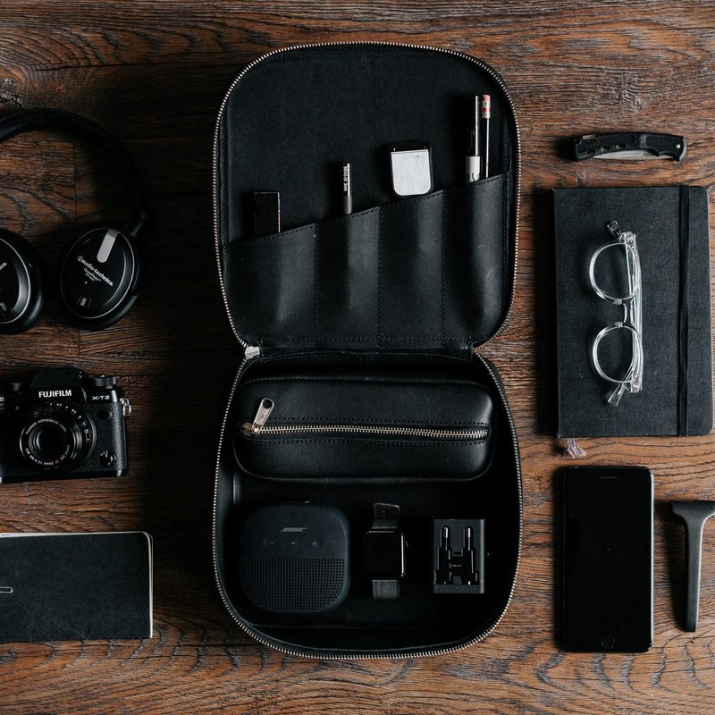 Flat-lay of all-black EDC (everyday carry) items, including headphones, a camera, glasses, a pocket knife, and more