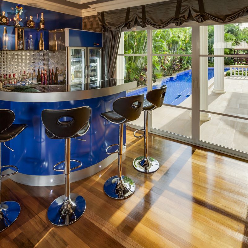 Home bar area with a blue-colored bar with bar stools next to floor to ceiling windows.