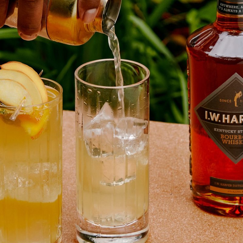 A Harper Spring Thyme being poured into a glass beside a glass of cocktail with fruit garnish and a bottle IW Harper Bourbon Whiskey.
