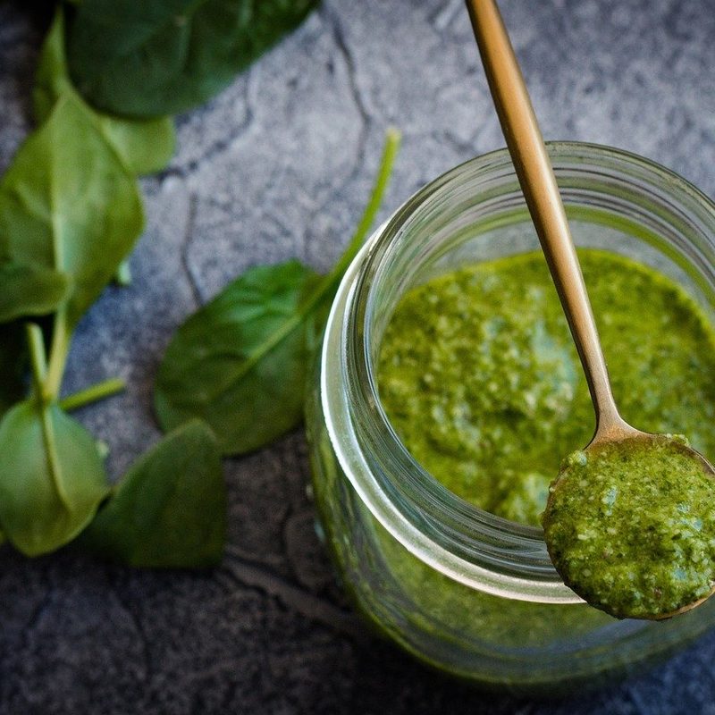 A long spoon holds fresh pesto above a jar of pesto.