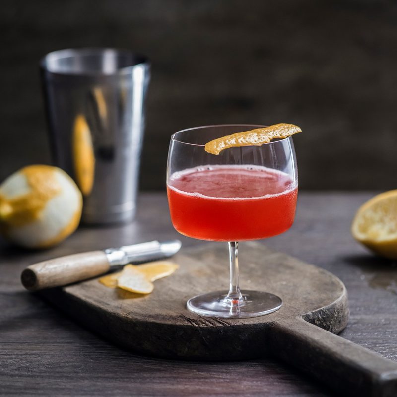 A Scofflaw in Love Cocktail with orange peel on top, served on a cutting board with a cocktail shaker and oranges in the background.