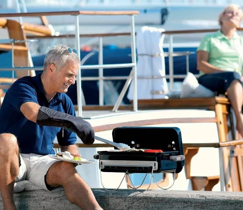 Man cooking on the Weber Go-Anywhere Charcoal Grill with a boat in the background.