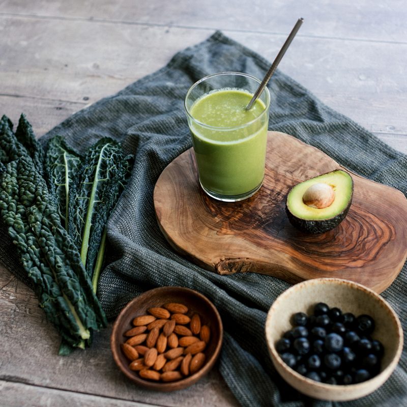 A glass of green smoothie with a metal drinking straw freshly served on a wooden board, with avocado, kale, almonds and blueberries on the table
