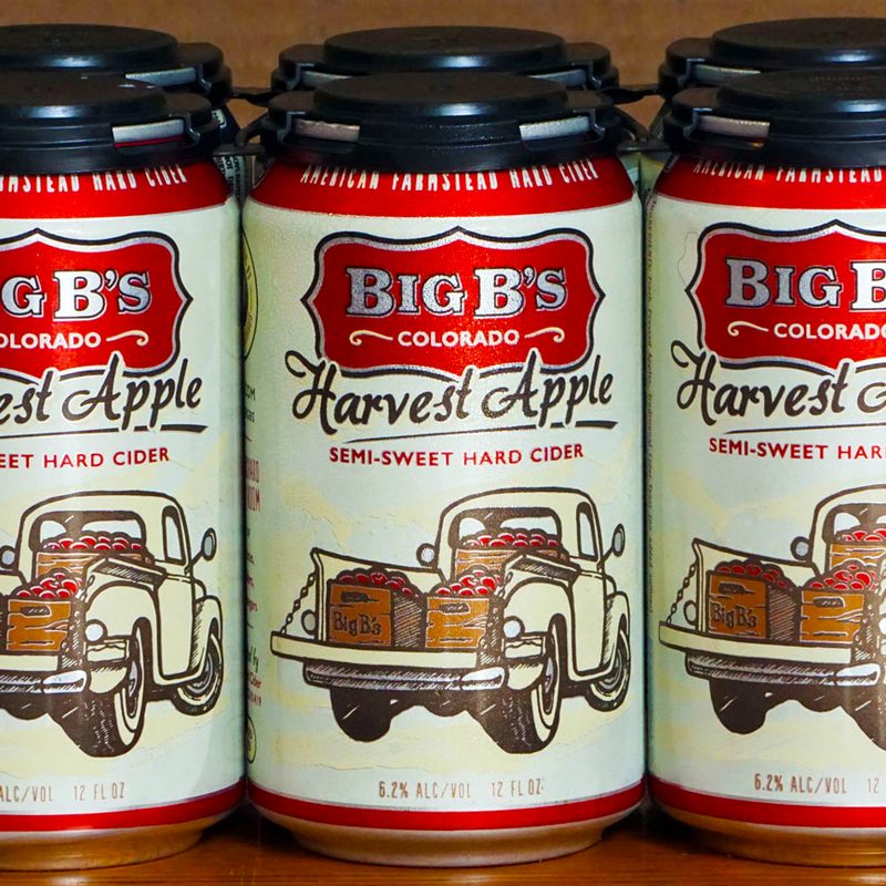 A six pack of Big B's Harvest Apple Hard Cider, lemon yellow cans with a vintage pickup loaded with apple crates.