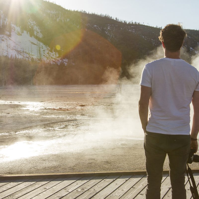 Rear view of a man standing with a digital camera at his side in front of a geyser at Yellowstone National Park.