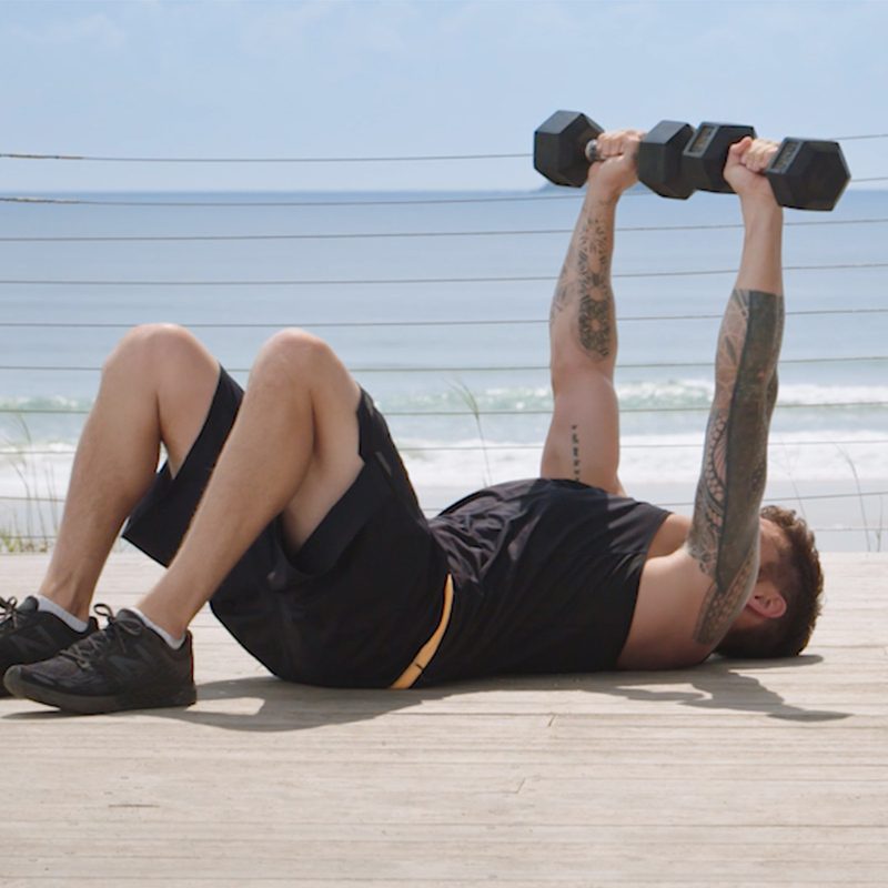 man in shorts at the beach laying on the ground doing chest presses with dumbbells.