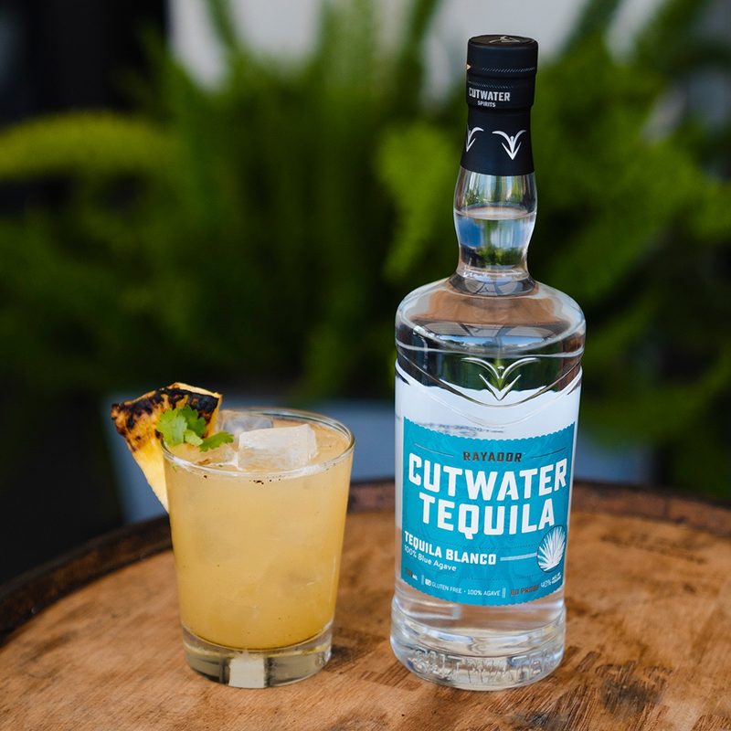 A bottle of Cutwater Tequila and a glass of Grill & Chill cocktail garnished with pineapple and leaves on a round wooden table