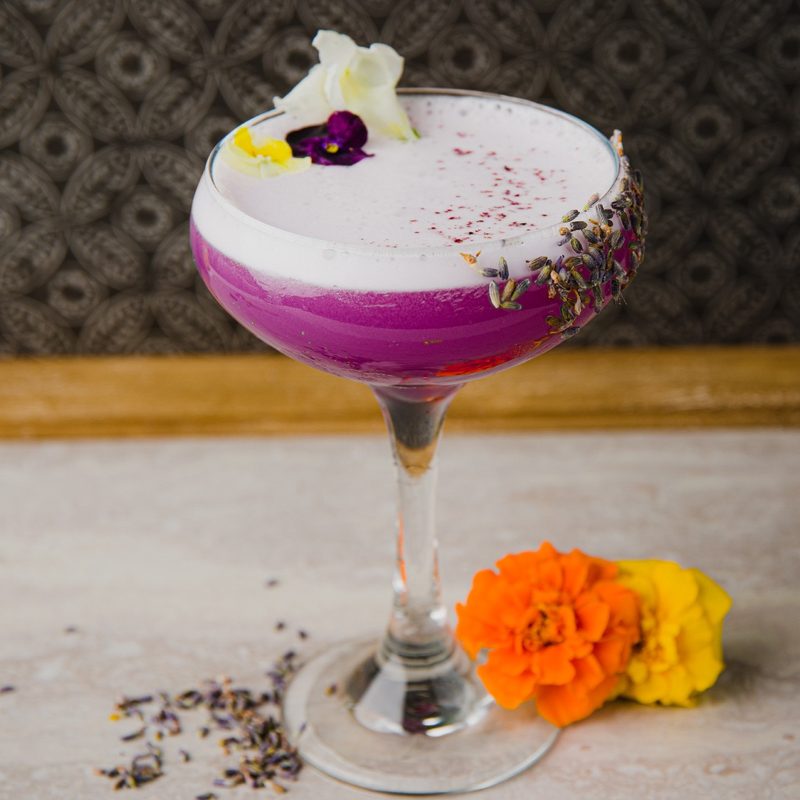 A Lavender Lasso cocktail with flowers and seeds beside it on a table