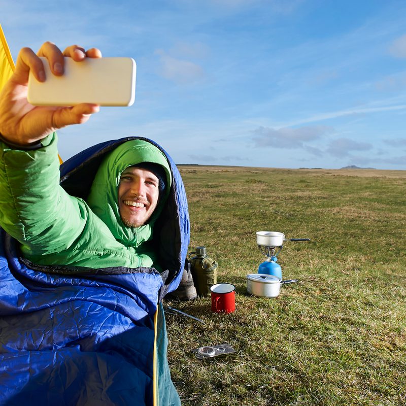 Camping man in a sleeping bag taking a selfie on a grass-covered hill.