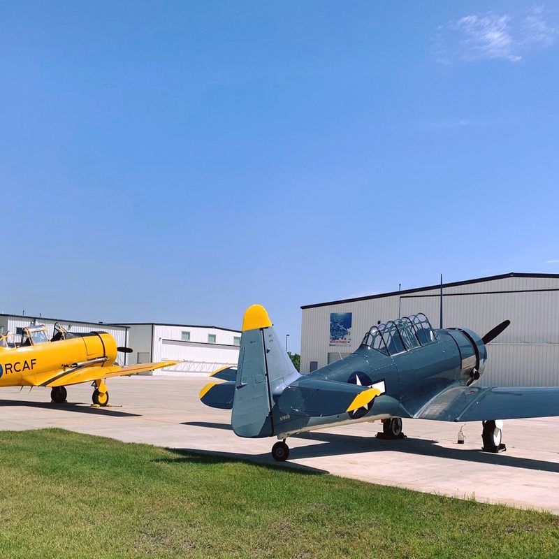 Two fighter planes, one yellow one black, sit facing an aircraft hangar.