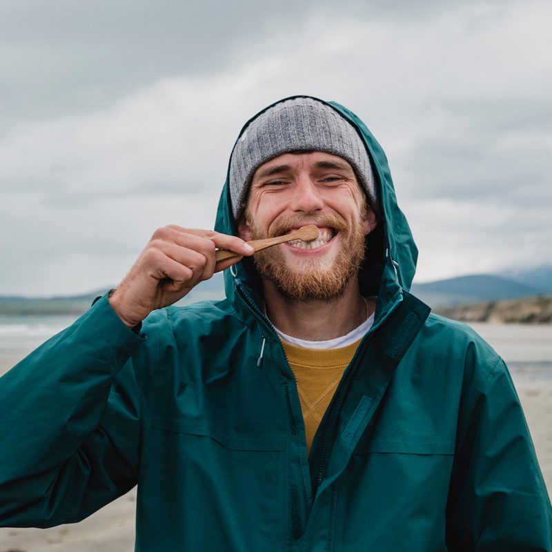 A young man brushing his teeth near a body of water, mountains in the background.