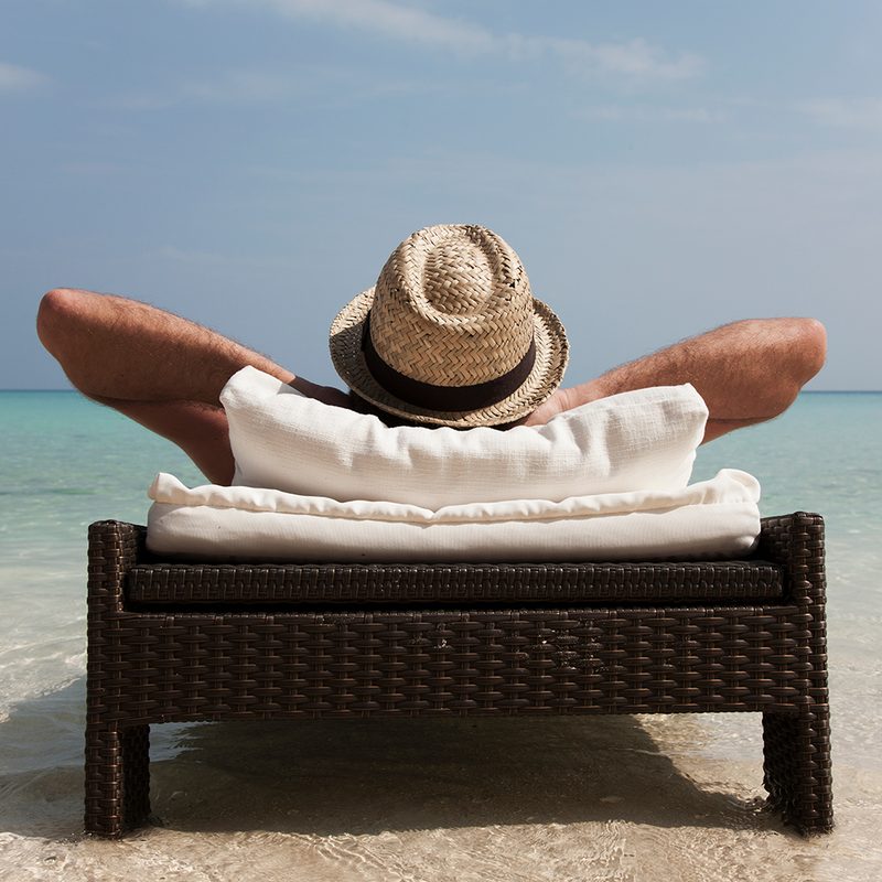 A man with his hands on the back of his head is wearing a straw hat and relaxing on the beach.