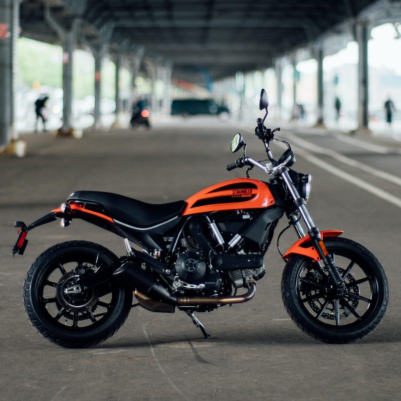 Ducati Scrambler Sixty2 motorcycle underneath a bridge with pedestrians and cars in the back