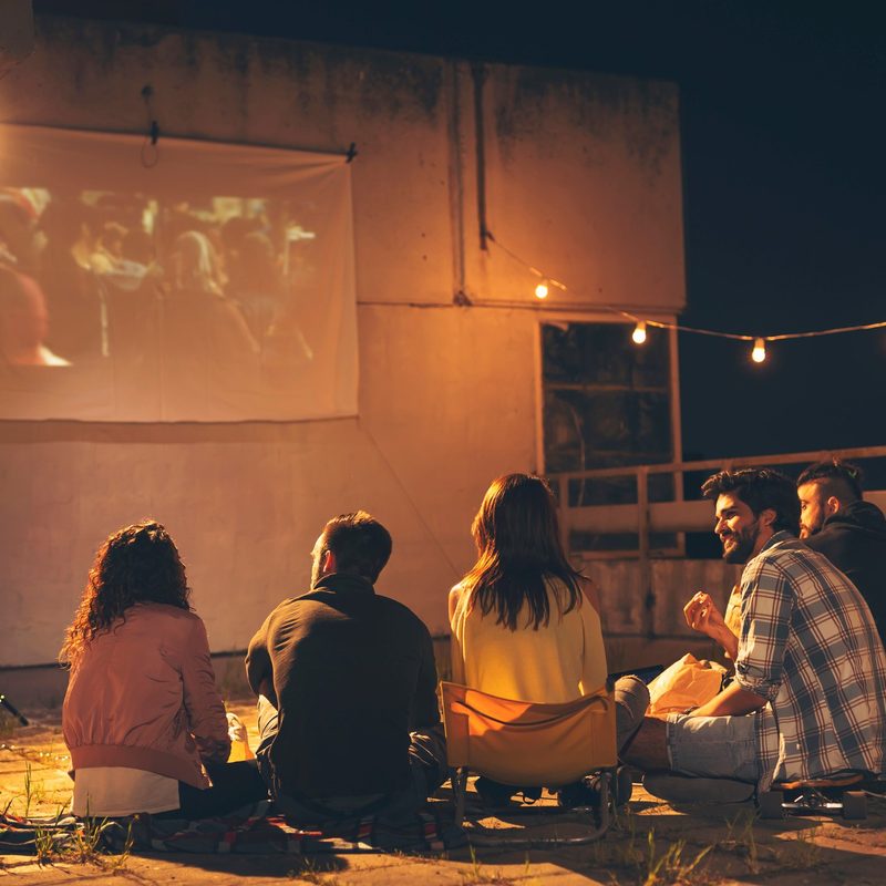 Group of young friends watching a movie on a building rooftop terrace, eating popcorn, drinking beer and having fun