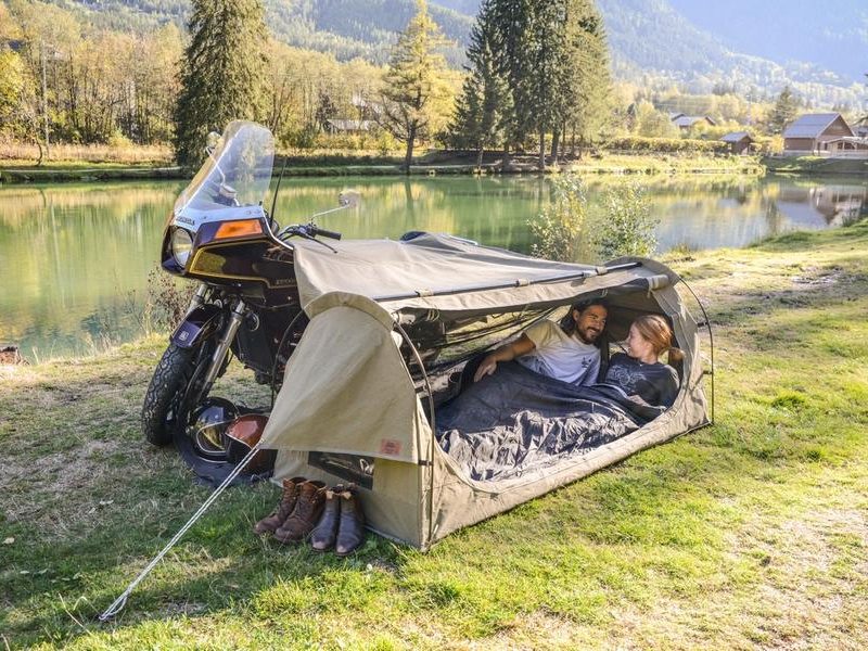 Two people sleeping inside a Wingman-of-the-Road Toucan Motorcycle Camping System beside a lake.