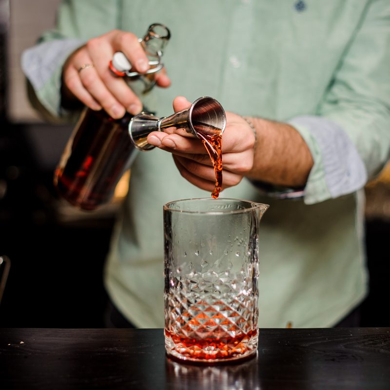 Bartender making alcoholic cocktail in red color, metal jigger and bar environment