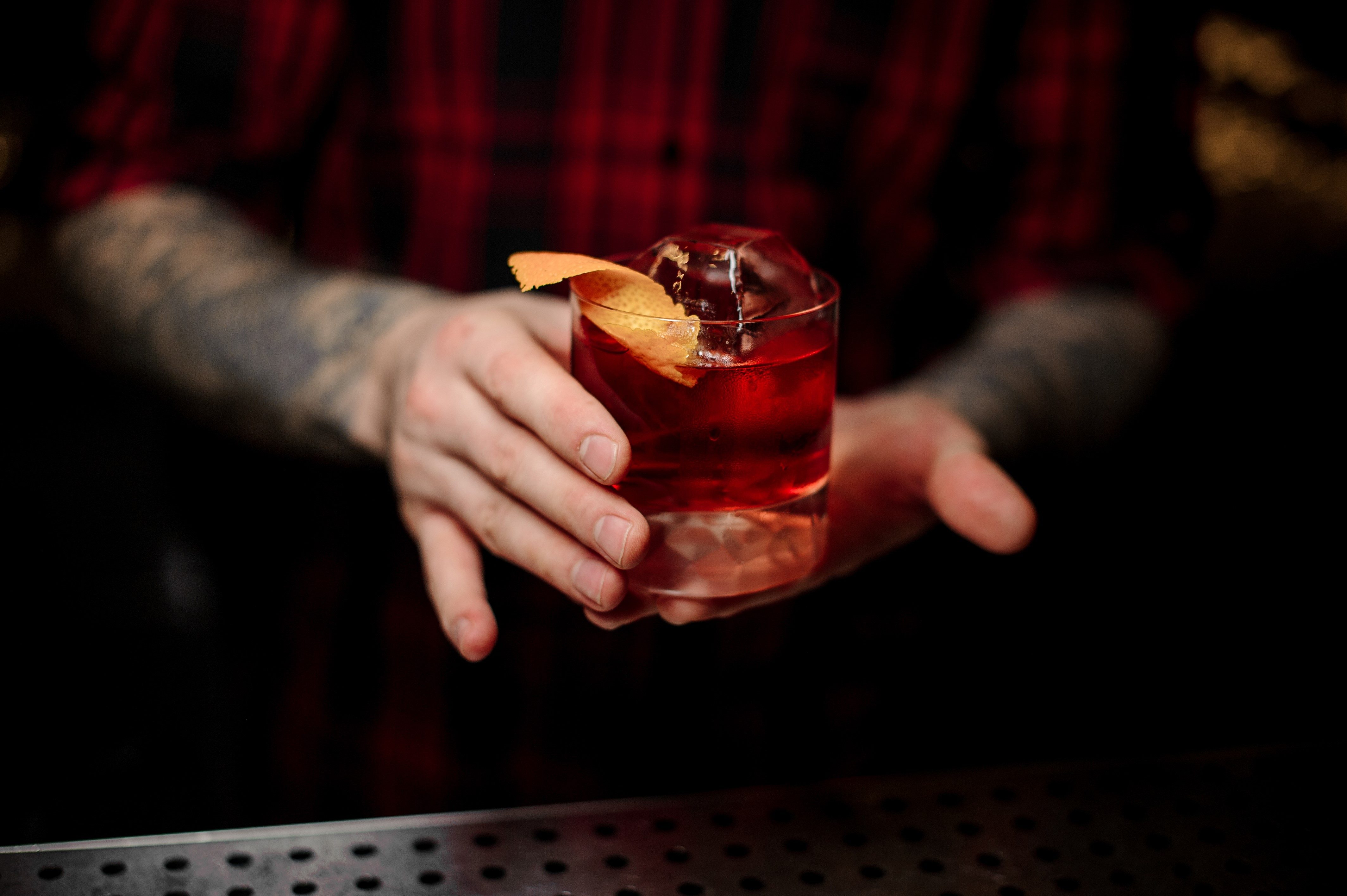 Bartender serving glass of a Boulevardier cocktail with big ice cube and orange zest on the bar counter on the blurred background