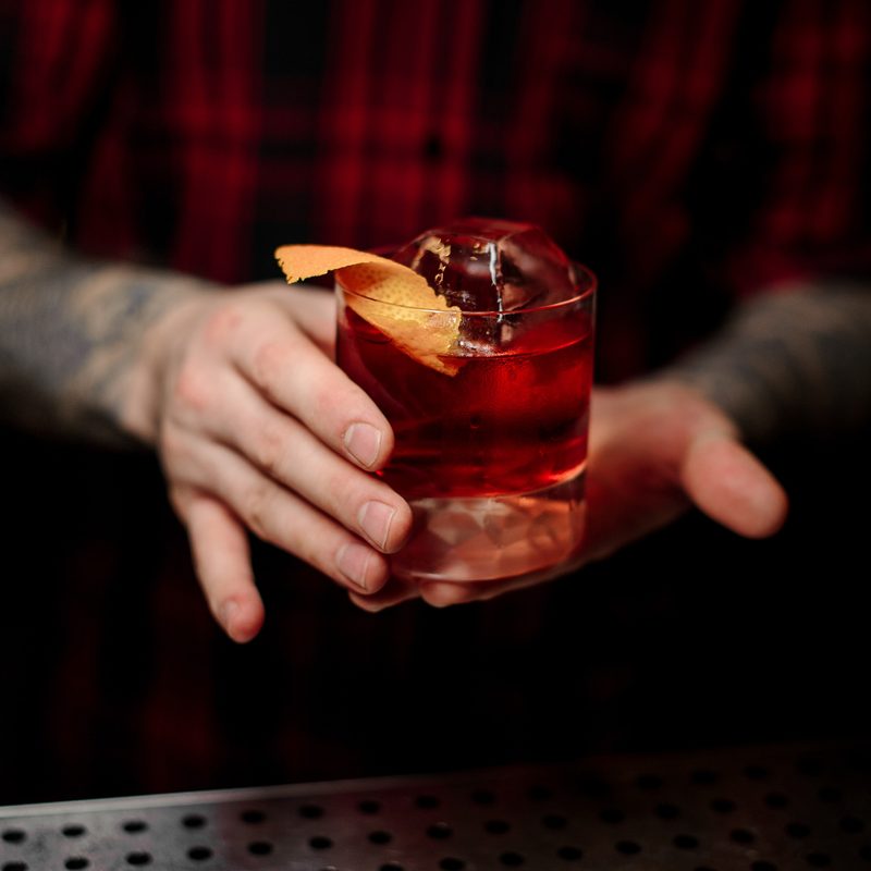 Bartender serving glass of a Boulevardier cocktail with big ice cube and orange zest on the bar counter on the blurred background