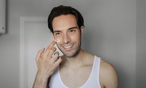 A Handsome Young Man Applying Moisturizing Cream on His Face While Standing in Front of the Mirror. Skin Care and Beauty Concept.