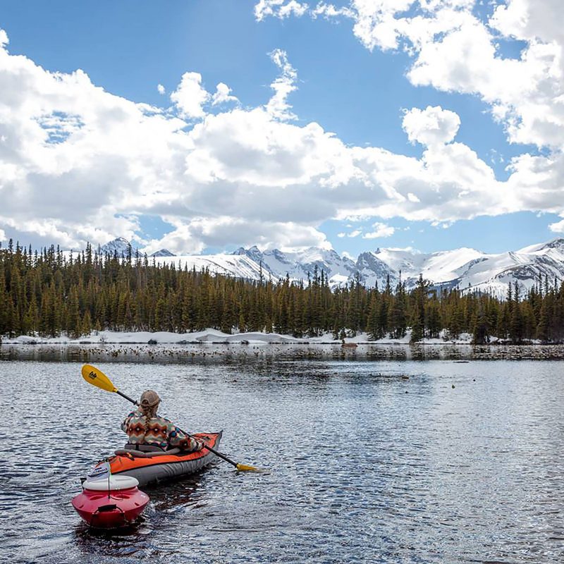 Kayaker paddling an inflatable kayak with a floating cooler in tow and snowy mountains in the background.