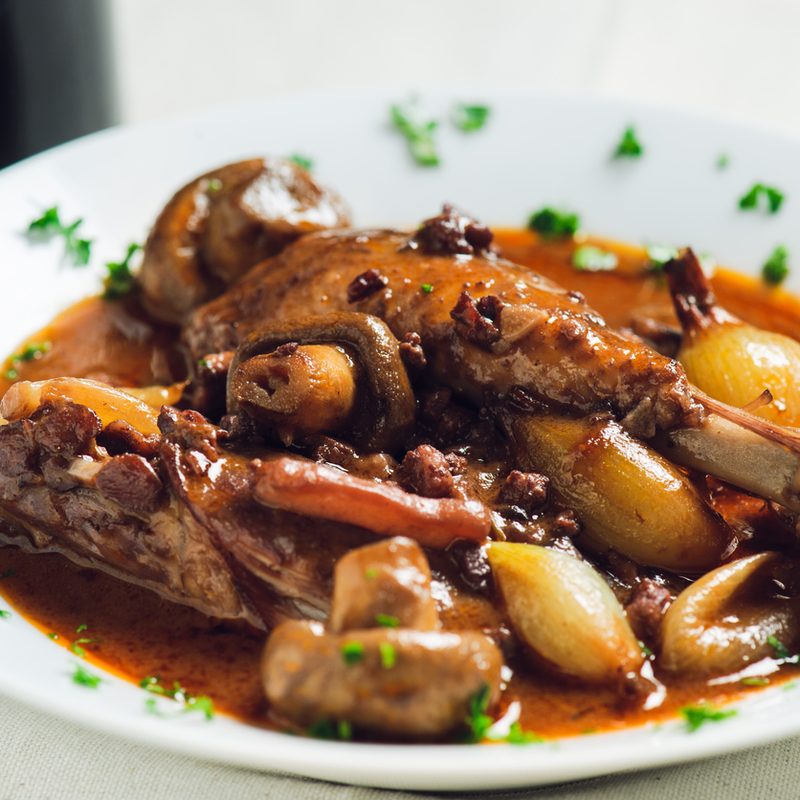 A plate of French traditional coq au vin -- chicken stew -- on a white table with bottle of wine in the frame.