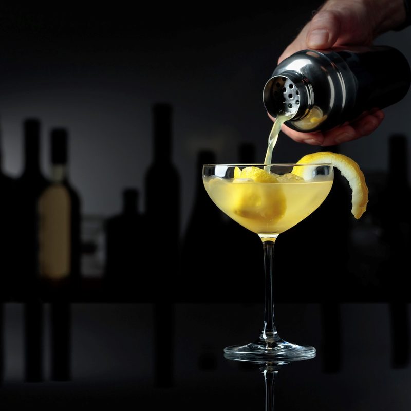 Cocktail "Sidecar" on a black reflective background. The bartender pours a cocktail from a shaker into a glass with ice