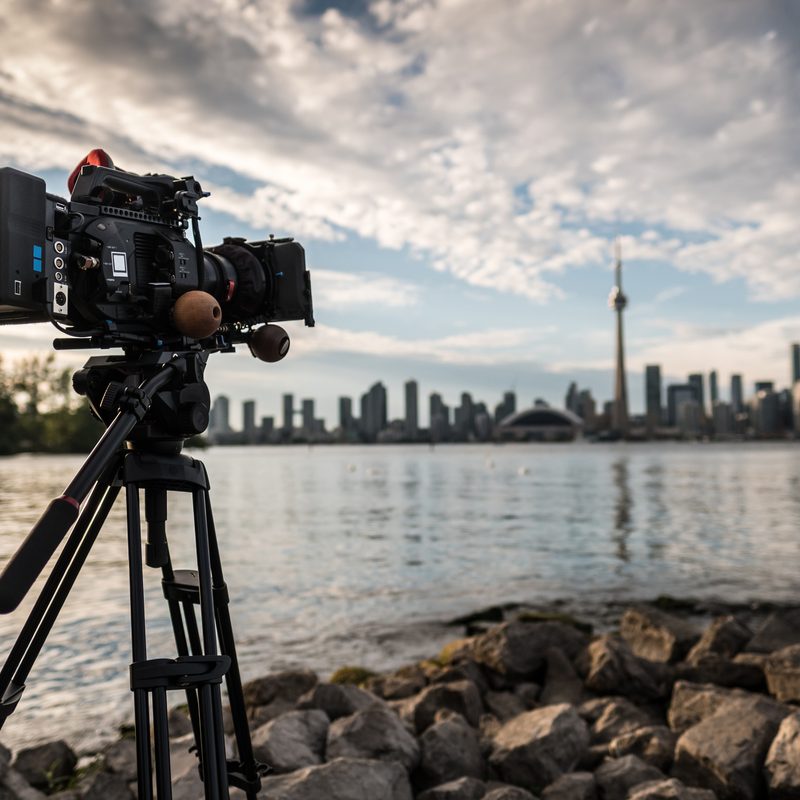 Toronto, Ontario, Canada. Professional 4K Video camera on tripod shooting time lapse of downtown panorama in sunset. Rocky beach of Toronto Island. Shallow depth of field with point of the focus on rear of the camera