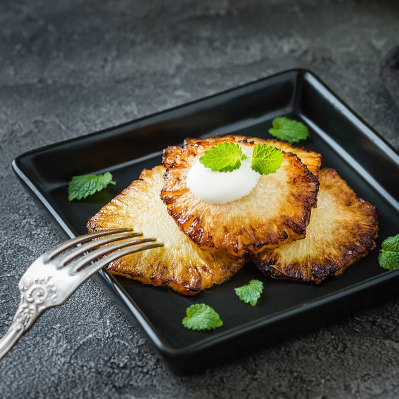 Fried pineapple slices with creme fraiche, mint in black square plate on dark background