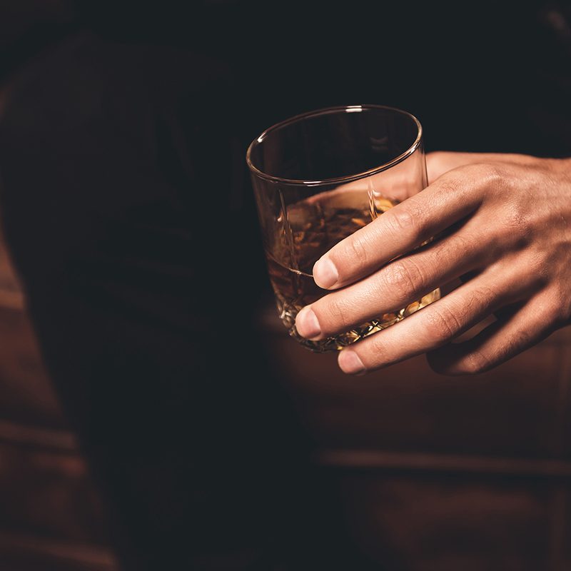 A close up of a man's hand holding a highball glass with about two fingers of whiskey in it.