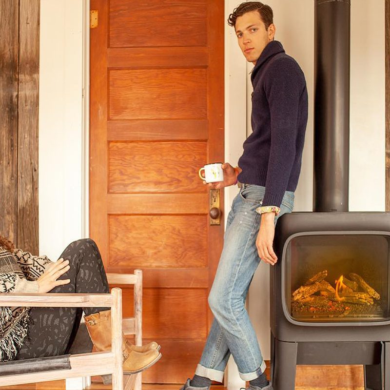A woman sitting on a chair with her legs up and a man with a cup standing close to a furnace.