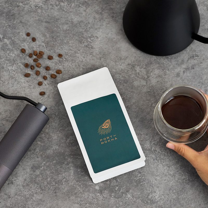 A bird’s eye shot of the Port of Mokha logo, coffee beans, assorted coffee essentials, and a hand holding a cup of coffee.