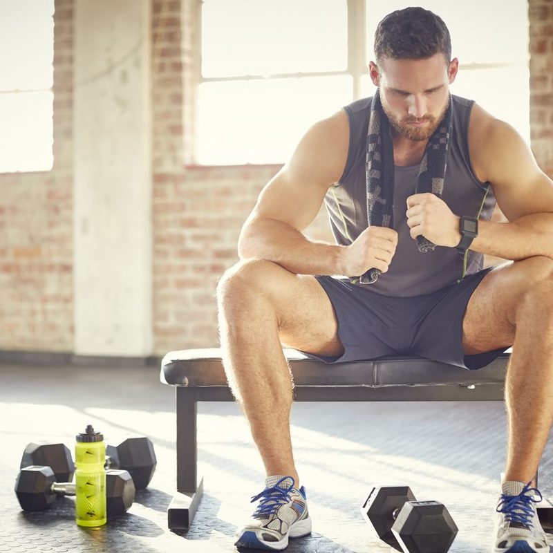 A tired man with towel sitting on a bench in a gym.