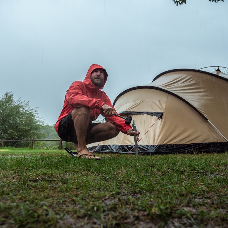 A man wearing a raincoat attempts to set up a tent in the rain.