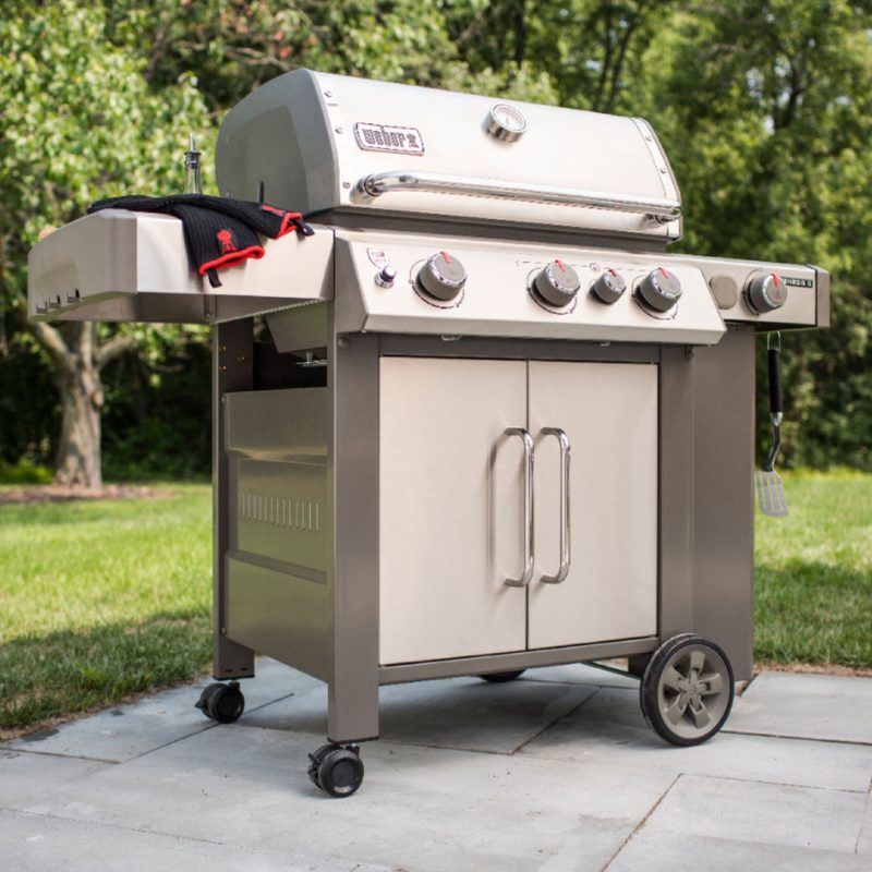 A Weber Genesis II S-335 3-burner propane gas grill on the edge of a patio with lawn in the background.