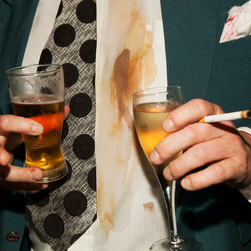 Midsection of a man holding drinks and cigarette at party with a beer stain on his shirt.