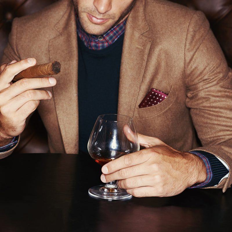 A stylish man drinking whiskey and smoking a cigar.