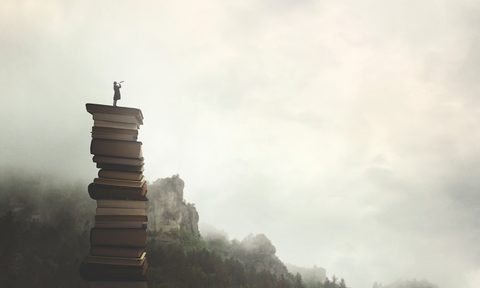 surreal man with telescope looks at infinity from the top of a stack of books in the outdoors.