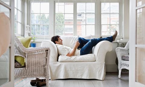 Man laying down on couch reading book next to window.